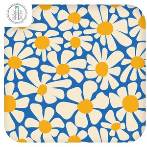 May include: A square textile featuring a repeating pattern of white daisy flowers with yellow centres against a vibrant blue background. The top left corner displays a circular logo with the text DELA BELA and the word home repeated around the edge.
