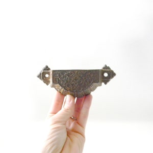 May include: A vintage cast iron drawer pull with an ornate design. The pull is a light brown color and has a slightly tarnished finish.