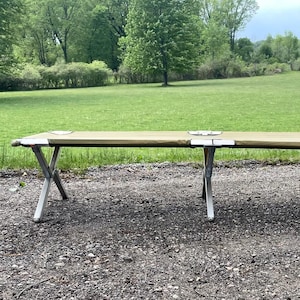 May include: A tan and silver folding cot set up outdoors on a gravel surface. The cot has a rectangular tan canvas top and silver metal legs. The background features a green grassy field and trees.