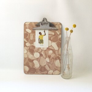 May include: A brown and white patterned clipboard with a metal clip. A small card with a drawing of a person waving is clipped to the clipboard. The card says "waving".