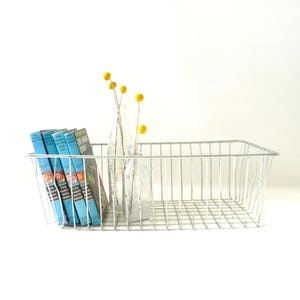 Puede incluir: Una cesta de alambre de metal blanco con un juego de libros de bolsillo azules y un jarrón de vidrio con flores secas amarillas.
