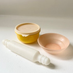 May include: A set of three kitchen tools: a yellow bowl with a lid, a pink bowl, and a white rolling pin.