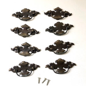 May include: Set of eight antique brass cabinet pulls with ornate designs. Each pull has a loop handle and comes with two screws.