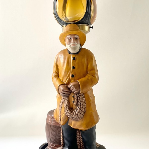 Può includere: Una lampada in ceramica a forma di pescatore che tiene una corda, in piedi su una botte con un impermeabile giallo e un cappello nero. La lampada ha un paralume in vetro con un design giallo e nero.
