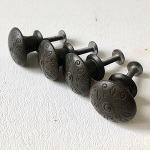 May include: Four dark brown metal cabinet knobs with a decorative floral design. The knobs have a round shape and a screw-on back.
