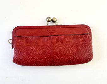 May include: A red leather Fossil wallet with a kiss-lock clasp. The wallet features an embossed geometric pattern and a small metal loop on the side. The Fossil logo is visible on the top. The clasp is made of two small, round, brass-colored balls.