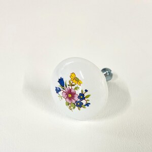 May include: A round white ceramic cabinet knob featuring a colorful floral pattern in the center. The design includes a large pink flower surrounded by smaller blue and yellow blossoms with green leaves. A silver metal screw extends from the back.