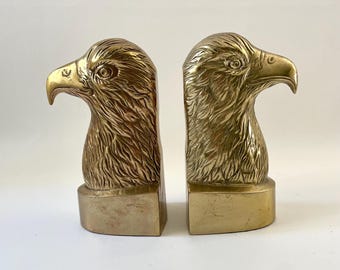 May include: A pair of brass eagle head bookends. Each bookend features a detailed sculpture of an eagle's head with a curved beak and textured feathers. The bookends have a rectangular base and a polished finish.