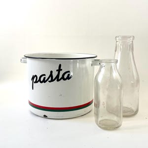 May include: A white enamel pasta pot with black lettering and red and green stripes, next to two clear glass bottles. The pot has two handles and a black rim. The bottles are different sizes.