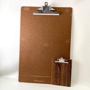 May include: Two clipboards, one large and one small, are displayed against a white background. The large clipboard is a solid brown color with a silver clip at the top. The smaller clipboard has a wood grain pattern and a silver clip.