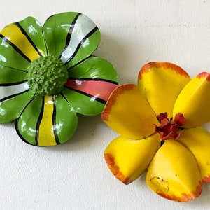 May include: Two decorative metal flower ornaments. One is green with yellow, white, and pink accents. The other is yellow with red-orange edges and a red center. Both are on a white surface.