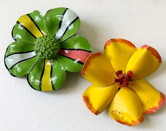 May include: Two decorative metal flower ornaments. One is green with yellow, white, and pink accents. The other is yellow with red-orange edges and a red center. Both are on a white surface.