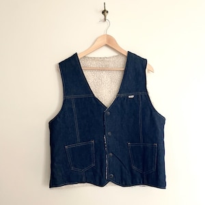 May include: A dark blue denim vest with a cream-colored, fuzzy lining. The vest has two front pockets, a V-neck, and a button-up closure. It is hanging on a wooden hanger against a white wall.