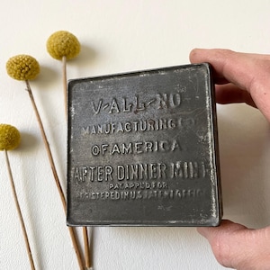 May include: A vintage metal tin with embossed text reading "V-ALL-NO MANUFACTURING OF AMERICA AFTER DINNER MINT". The tin is rectangular, with a dark, aged appearance. Yellow dried flowers are in the background.