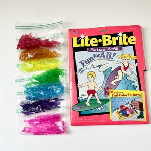 May include: Lite-Brite Picture Refill box with a pink background. The box features a picture of two children on a surfboard. The box includes 12 full-color pictures and 8 guide sheets to create your own designs. The box also includes a bag of rainbow-colored pegs.