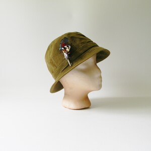 May include: A green felt cloche hat with a feather accent. The hat is on a white mannequin head.