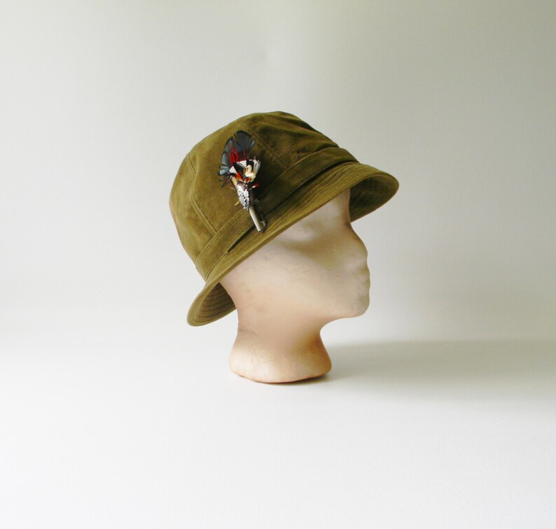 May include: A green felt cloche hat with a feather accent. The hat is on a white mannequin head.