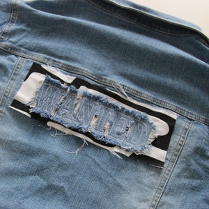 Peut inclure: Veste en jean bleu clair avec un patch "WANTED" vieilli au dos. Le patch est en jean et présente des bords effilochés, sur un tissu à rayures noires et blanches.