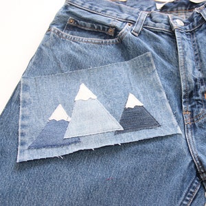 May include: A pair of blue denim jeans with a patch of denim sewn on the front pocket. The patch is shaped like three mountains with white peaks.