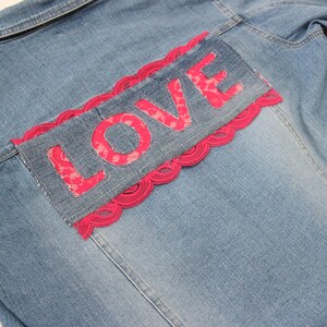 Puede incluir: Una chaqueta vaquera azul con un ribete de encaje rosa y la palabra "LOVE" cosida en la espalda.
