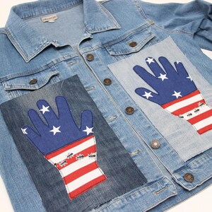 Puede incluir: Una chaqueta vaquera azul con dos parches cosidos. Cada parche es una pieza de tela en forma de mano con un diseño de bandera estadounidense roja, blanca y azul. Los parches tienen pequeñas hormigas negras cosidas en ellos.