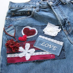 Op de afbeelding: Blauwe denim jeans met verschillende stoffen patches, waaronder rode harten, witte kant en een patch met de tekst "LOVE" in witte borduurwerk.