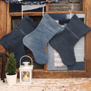 May include: Three blue denim Christmas stockings hanging on a rustic wooden window frame. The stockings are made from repurposed denim fabric and have a patchwork design. The window frame is decorated with more denim fabric scraps.