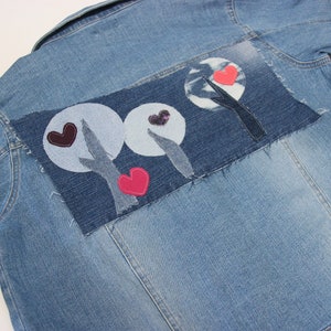 May include: A blue denim jacket with a patchwork design on the back. The design features three circles with hearts and trees in a stylized design.