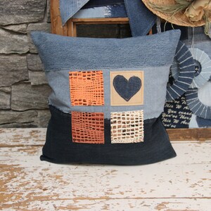 Puede incluir: Una almohada de mezclilla azul con un diseño de patchwork. La almohada tiene cuatro cuadrados de tela naranja y beige con una pieza de mezclilla en forma de corazón en el centro.