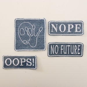 Op de afbeelding: Vier denim patches met wit geborduurde ontwerpen en tekst. Eén patch toont een alienachtig gezicht, terwijl de andere "NOPE", "NO FUTURE" en "OOPS!" lezen. De patches hebben een rechthoekige vorm met afgeronde hoeken.