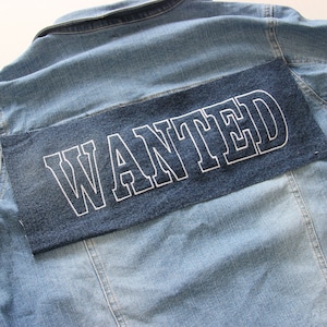 Puede incluir: Una chaqueta vaquera azul con un parche de mezclilla azul oscuro cosido en la espalda. El parche tiene la palabra "WANTED" bordada en blanco.