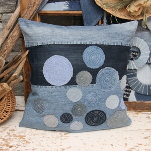 May include: A blue denim pillow with a patchwork design featuring circles of different sizes and shades of blue. The pillow is decorated with a variety of denim scraps, creating a unique and textured look.