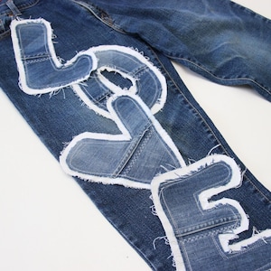 May include: A pair of blue denim jeans with the word "LOVE" spelled out in white fabric patches. Each letter is made from a different piece of denim.
