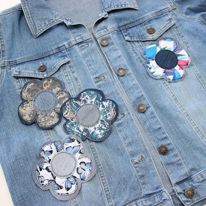 Può includere: Una giacca di jeans blu chiaro con quattro toppe di fiori in tessuto. I fiori sono di vari colori e motivi, tra cui blu, bianco, rosso e verde.