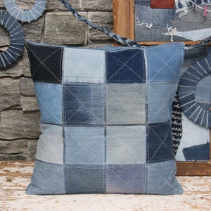Puede incluir: Una almohada cuadrada de mezclilla con un diseño de patchwork. La almohada está hecha de diferentes tonos de mezclilla azul y está cosida con hilo blanco.