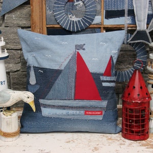 May include: A blue and white lighthouse figurine with the text "NEW PORT" on the side. A white wooden seagull figurine is perched on a wooden barrel. A red metal lantern with a cage design is next to a white wooden lighthouse figurine. A blue denim pillow with a red sailboat design and the text "GLORIA UNLIMITED" is in the centre of the image.