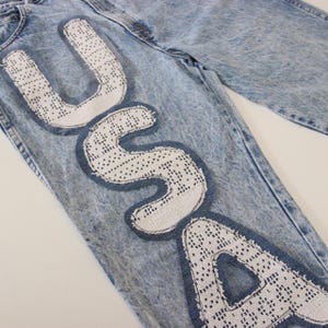 Könnte beinhalten: Hellblaue Jeans im Used-Look mit den Buchstaben "USA" auf der Vorderseite. Die Buchstaben sind weiß, aus spitzenähnlichem Material und mit dunklerem blauem Denim umrandet.