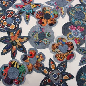 May include: A collection of denim flower appliqués with various patterned fabric centers. The flowers are in different shapes and sizes, with some featuring heart-shaped accents. The color palette includes shades of blue, red, orange, and yellow.