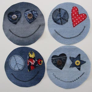 Op de afbeelding: Vier glimlachende gezichten van denim stof met verschillende versieringen. Eén heeft twee harten met knopen als ogen, één heeft een vredesteken en een rood hart met witte stippen, één heeft een vredesteken en een bloemig hart, en één heeft een hart met een knoop en een ster met een knoop.