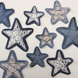 May include: A selection of denim star-shaped patches in different sizes. Some stars have white lace or printed text on a grey background, while others are plain denim. The patches are displayed on a white surface.