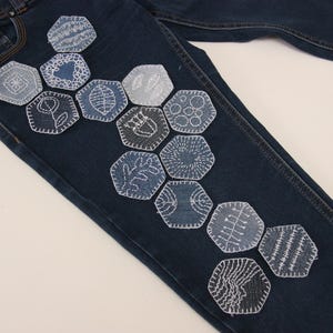 May include: Dark blue denim jeans with a series of embroidered, hexagon-shaped patches in various shades of blue and white. The patches feature different designs, including floral, geometric, and abstract patterns. The jeans are shown on a white surface.
