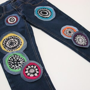 Peut inclure: Un jean bleu denim avec des patchs en tissu colorés cousus dessus. Les patchs sont circulaires et présentent une variété de motifs floraux.