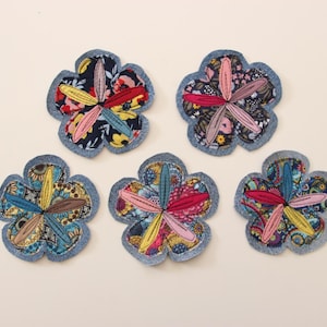 Op de afbeelding: Vijf handgemaakte bloemvormige stoffen onderzetters. Elke onderzetter heeft een denimblauwe rand en een kleurrijk bloemenpatroon in het midden. De onderzetters zijn gerangschikt op een crèmekleurig oppervlak.