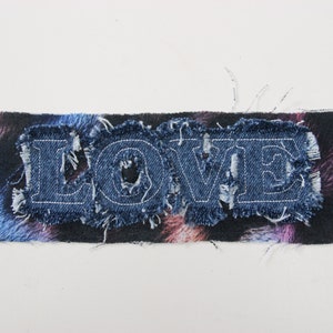 Op de afbeelding: Een denim patch met het woord "LOVE" in witte stiksels op een zwarte achtergrond met een abstract patroon in blauw, roze en paars.