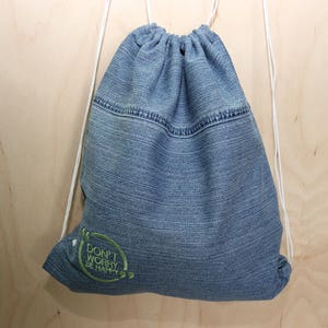 Op de afbeelding: Een blauwe denim trekkoordtas met witte trekkoorden. De tas heeft een groene geborduurde quote die luidt "Maak je geen zorgen, wees gelukkig".