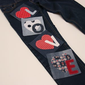 Puede incluir: Vaqueros azules de denim con corazones de tela roja, blanca y negra cosidos sobre ellos. Los corazones deletrean la palabra "LOVE" en letras mayúsculas.