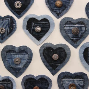 Puede incluir: Una colección de 12 adornos en forma de corazón de mezclilla con botones de metal en el centro. Los corazones están hechos de diferentes tonos de mezclilla azul y están dispuestos en un patrón aleatorio.
