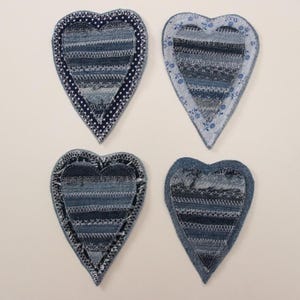 Op de afbeelding: Vier hartvormige denim patches met verschillende blauwe stofpatronen en stiksels. Elk hart heeft een uniek randontwerp, inclusief stippen en bloemmotieven. De patches zijn gerangschikt op een lichte achtergrond.