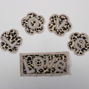 Pode incluir: Cinco apliques de tecido com um design de estampa de leopardo. Quatro são em forma de flor, soletrando "LOVE" com letras bege. O quinto é um patch retangular com a palavra "LOVE" no mesmo estilo. Os apliques estão em um fundo branco.