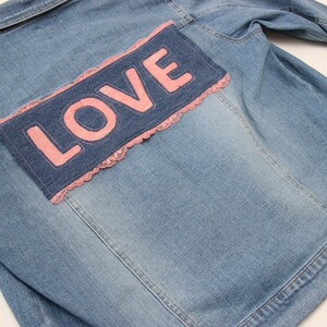 May include: A light blue denim jacket with a pink "LOVE" patch on the back. The patch is surrounded by a white lace trim.